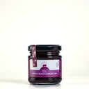 A glass jar of The Mulberry Pigs Apple & Black Currant Jam with a black lid and a purple label, set against a plain light gray background.
