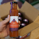 A hand holds a glass bottle of The Mulberry Pig Tomato Ketchup with cloves and bay leaves, with an open cardboard box of more bottles in the background.