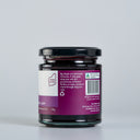 A glass jar of The Mulberry Pig Apple and Blackcurrant jam showing the back of the label with product details, set against a clean light gray background.