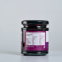 A glass jar of The Mulberry Pigs Apple & Black Currant Jam with a black lid and a purple label showing nutritional information, set against a plain light gray background.