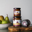 Two glass jars of jam with black lids are arranged on a wooden board against a light gray background. One jar is stacked on top of the other, and a third jar lies on its side.