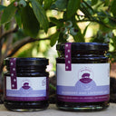 Two glass jars of Blueberry and Lavender Jam by The Mulberry Pig, one small and one large, with purple labels and black lids, set outdoors in front of a leafy bush.