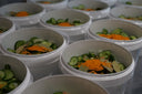 Several white plastic tubs filled with sliced cucumbers and bright orange turmeric spice powder, arranged closely together on a dark surface. ready to become the mulberry pigs bread and butter pickles
