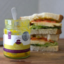 A jar of piccalilli open in front of a Cheese Sandwich on a wooden chopping board