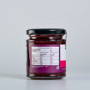 A glass jar of The Mulberry Pig Raspberry Jam  with a black lid, showing the back of the label with nutrition information. The jar is set against a clean, light gray background.