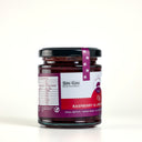 A glass jar of Raspberry & Lime Jam with Gin by The Mulberry Pig, featuring a white and red label with a black lid and a purple seal ribbon, set against a plain light gray background.
