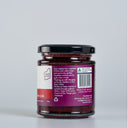 A glass jar of The Mulberry Pig Raspberry & Lime Jam with Gin, showing the back of the label with product details, set against a clean light gray background.