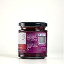 A glass jar of The Mulberry Pig Raspberry & Lime Jam with Gin, showing the back of the label with product details, set against a clean light gray background.
