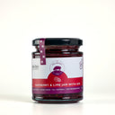A glass jar of Raspberry & Lime Jam with Gin by The Mulberry Pig, featuring a white and red label with a black lid and a purple seal ribbon, set against a plain light gray background.