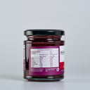 A glass jar of The Mulberry Pig, Raspberry & Lime Jam with Gin with a black lid, showing the back of the label with nutrition information. The jar is set against a clean, light gray background.
