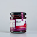 A glass jar of Raspberry & Lime Jam with Gin by The Mulberry Pig, featuring a white and red label with a black lid and a purple seal ribbon, set against a plain light gray background.