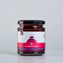 A glass jar of Raspberry Jam by The Mulberry Pig, featuring a white and magenta label , sealed with a black lid and a matching ribbon tag, set against a light gray background.