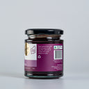A glass jar of Spiced Plum Butter with a black lid, showing the back of the label with description. The jar is set against a clean, light gray background.