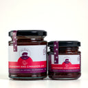 Two glass jars of The Mulberry Pig Strawberry and Cinnamon Jam on a clean white background, one large jar and one smaller jar, both with black lids and white labels featuring a red strawberry graphic.