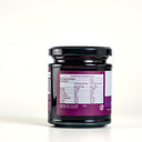 A glass jar of The Mulberry Pigs Apple & Black Currant Jam with a black lid and a purple label showing nutritional information, set against a plain light gray background.