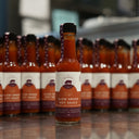 A bottle of Slow Smoke Hot Sauce stands in sharp focus on a reflective countertop, with multiple identical bottles blurred in the background.