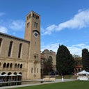 A tall stone clock tower on a historic building beside a reflecting pool, with a manicured green lawn in the foreground and a bright blue sky with white clouds overhead.
