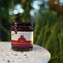 A glass jar of The Mulberry pig Raspberry & Lime Jam with Gin sits on a white metal patio table with a blurred garden background.