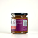 A glass jar of The Mulberry Pig Peach, Chilli and Black Pepper Chutney on a clean white background. The jar has a black lid, a purple and white label showing product description, and a purple seal ribbon, with visible chunks of reddish-brown chutney inside.