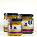 Three glass jars of Piccalilli from The Mulberry Pig, arranged in a triangular composition on a clean white background. The jars contain a yellow-green spread and are topped with black lids, featuring white and purple labels with a pig logo.