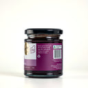 A glass jar of Spiced Plum Butter with a black lid, showing the back of the label with description. The jar is set against a clean, light gray background.