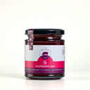 A glass jar of Raspberry Jam by The Mulberry Pig, featuring a white and magenta label , sealed with a black lid and a matching ribbon tag, set against a light gray background.