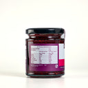 A glass jar of The Mulberry Pig Raspberry Jam  with a black lid, showing the back of the label with nutrition information. The jar is set against a clean, light gray background.
