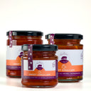 Three glass jars of Sweet Chilli Jam from The Mulberry Pig, arranged in a triangular composition on a clean white background. The front jar is in sharp focus, showing the dark red chili jam inside and its white and orange label with a purple brand ribbon.